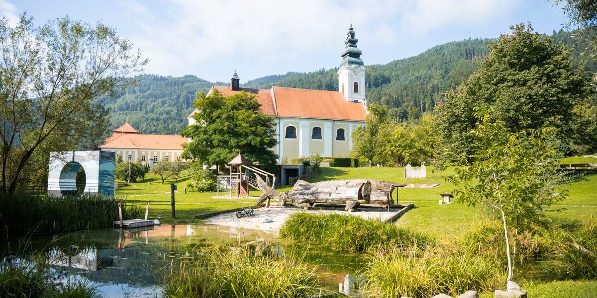 Eine weiße Kirche mit einem hohen Kirchturm steht neben einem Teich und grünen Rasenflächen, umgeben von Bäumen und Hügeln unter einem teilweise bewölkten Himmel.