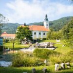 Eine weiße Kirche mit einem hohen Kirchturm steht neben einem Teich und grünen Rasenflächen, umgeben von Bäumen und Hügeln unter einem teilweise bewölkten Himmel.