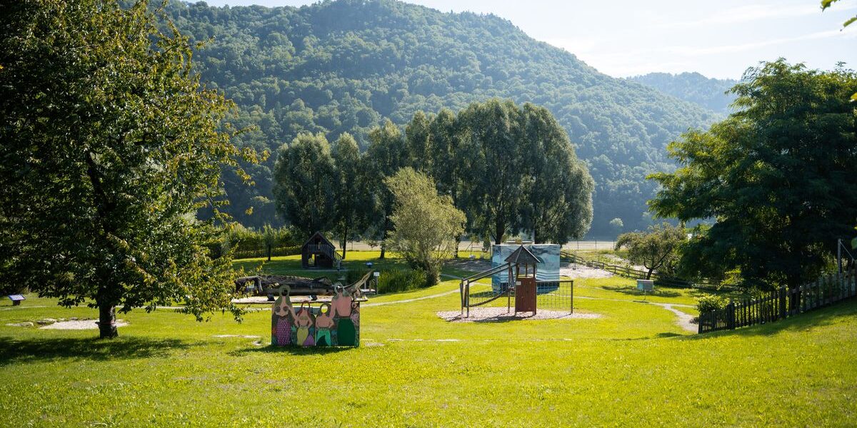 Ein grasbewachsener Park mit Spielgeräten, Bäumen und einem Berg im Hintergrund unter einem teilweise bewölkten Himmel.