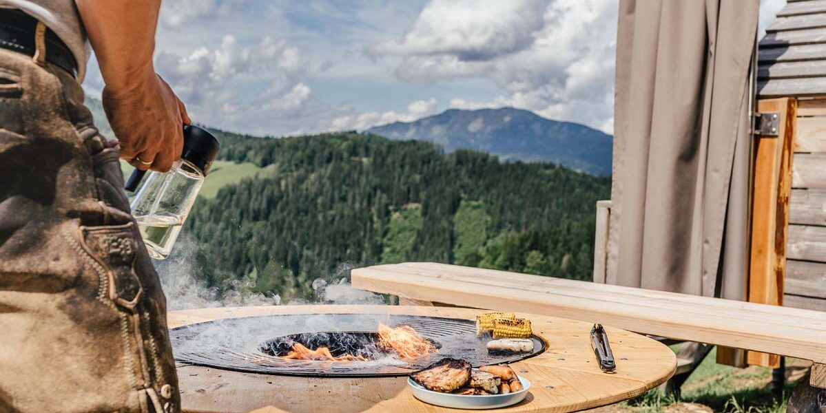 Eine Person hält einen Glaskrug neben einem Grill am Feuerplatz, auf dem Gemüse und Fleisch liegen; die Szene spielt im Freien vor dem Hintergrund von Bergen und Bäumen.