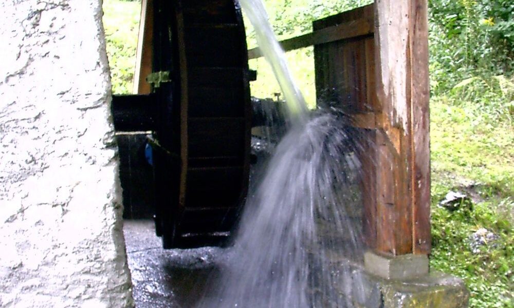 Ein hölzernes Wasserrad dreht sich, während das Wasser darüber fließt, und ist an einer Struktur aus Stein und Holz befestigt, die von Grün umgeben ist.