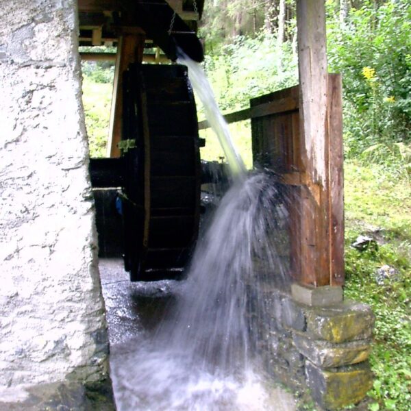 Ein hölzernes Wasserrad dreht sich, während das Wasser darüber fließt, und ist an einer Struktur aus Stein und Holz befestigt, die von Grün umgeben ist.