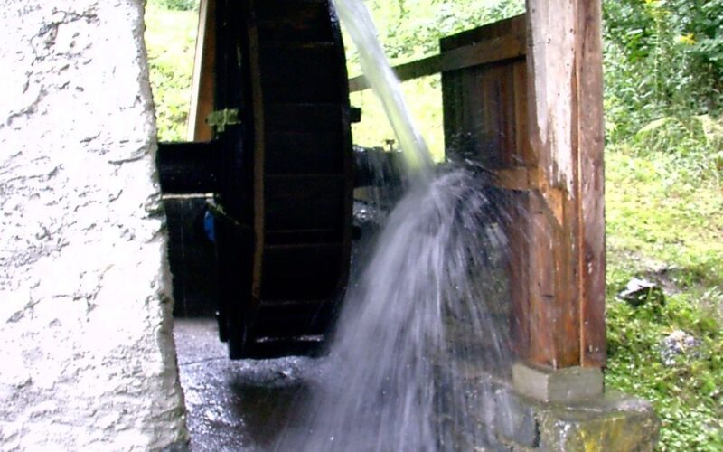Ein hölzernes Wasserrad dreht sich, während das Wasser darüber fließt, und ist an einer Struktur aus Stein und Holz befestigt, die von Grün umgeben ist.