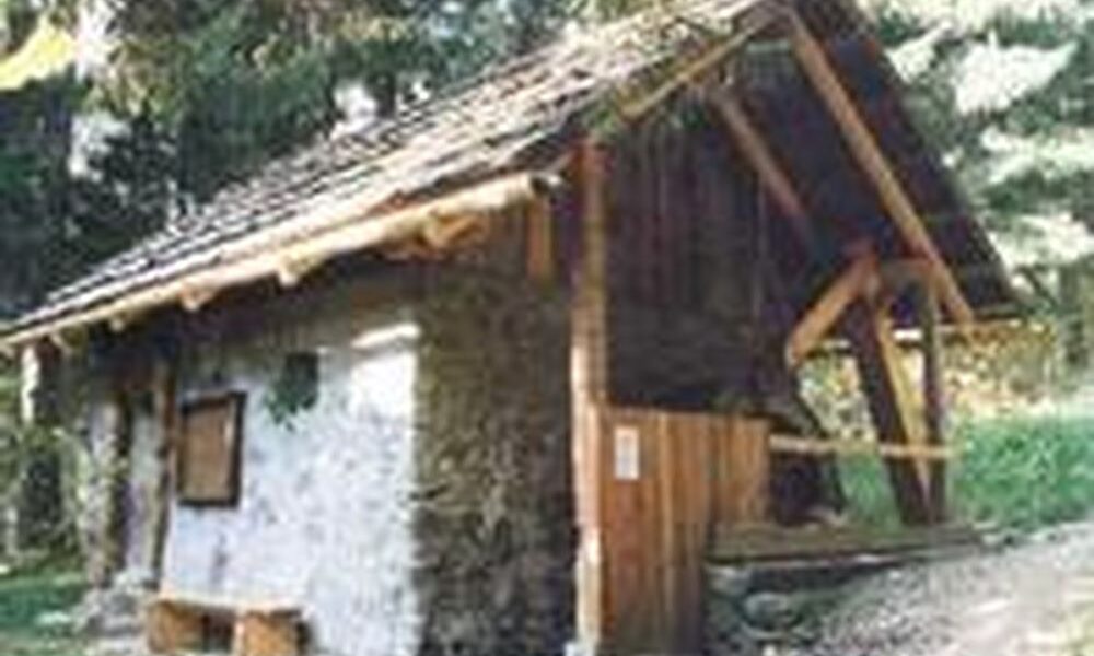 Eine kleine rustikale Hütte aus Stein und Holz mit schrägem Dach, umgeben von Bäumen, in einem Waldgebiet.