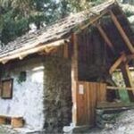 Eine kleine rustikale Hütte aus Stein und Holz mit schrägem Dach, umgeben von Bäumen, in einem Waldgebiet.