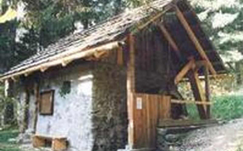 Eine kleine rustikale Hütte aus Stein und Holz mit schrägem Dach, umgeben von Bäumen, in einem Waldgebiet.