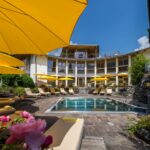 Außenpool mit gelben Sonnenschirmen und Liegestühlen, umgeben von einem geschwungenen, modernen Gebäude und an sonnigen Tagen von einer Gartenanlage.