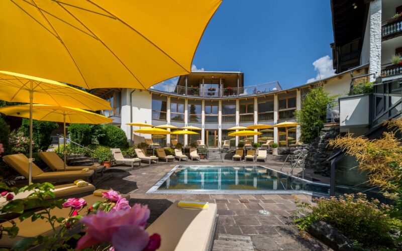 Außenpool mit gelben Sonnenschirmen und Liegestühlen, umgeben von einem geschwungenen, modernen Gebäude und an sonnigen Tagen von einer Gartenanlage.