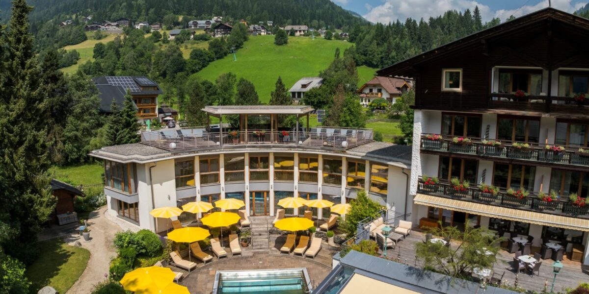Ein Hotel mit gelben Sonnenschirmen und einem kleinen Außenpool liegt in einer grünen, hügeligen Landschaft mit verstreuten Häusern und bewaldeten Flächen unter einem teilweise bewölkten Himmel.