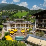 Ein Hotel mit gelben Sonnenschirmen und einem kleinen Außenpool liegt in einer grünen, hügeligen Landschaft mit verstreuten Häusern und bewaldeten Flächen unter einem teilweise bewölkten Himmel.
