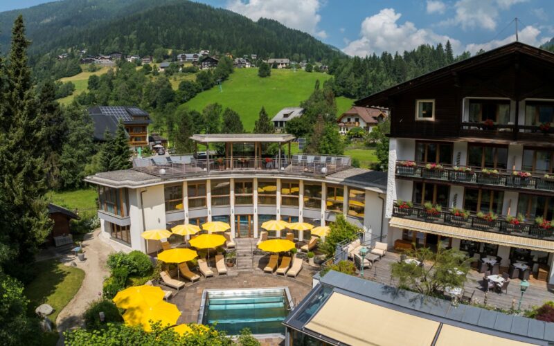 Ein Hotel mit gelben Sonnenschirmen und einem kleinen Außenpool liegt in einer grünen, hügeligen Landschaft mit verstreuten Häusern und bewaldeten Flächen unter einem teilweise bewölkten Himmel.