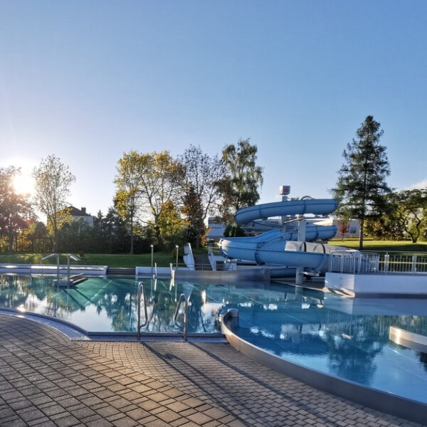 Freibad mit blauer Spiralrutsche, umgeben von Bäumen und Grün, unter einem klaren Himmel mit tiefstehender Sonne am Horizont.