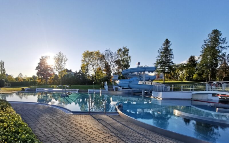 Freibad mit blauer Spiralrutsche, umgeben von Bäumen und Grün, unter einem klaren Himmel mit tiefstehender Sonne am Horizont.