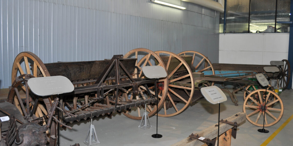 In der Halle sind mehrere antike Landmaschinen aus Holz und Metall mit Rädern ausgestellt, die jeweils mit einer Informationstafel versehen sind.