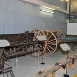 In der Halle sind mehrere antike Landmaschinen aus Holz und Metall mit Rädern ausgestellt, die jeweils mit einer Informationstafel versehen sind.