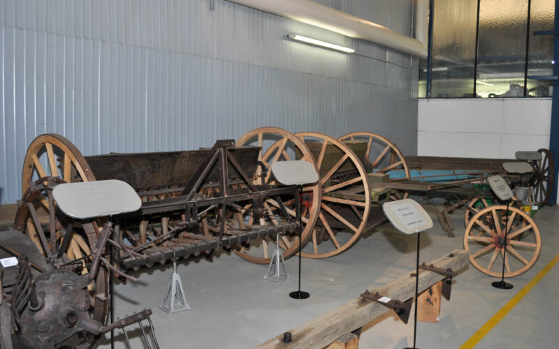 In der Halle sind mehrere antike Landmaschinen aus Holz und Metall mit Rädern ausgestellt, die jeweils mit einer Informationstafel versehen sind.
