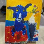 Ein farbenfrohes, abstraktes Gemälde eines blauen Hundes mit gelben Augen auf rotem und blauem Hintergrund, ausgestellt auf einer kleinen Staffelei.