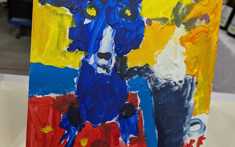 Ein farbenfrohes, abstraktes Gemälde eines blauen Hundes mit gelben Augen auf rotem und blauem Hintergrund, ausgestellt auf einer kleinen Staffelei.