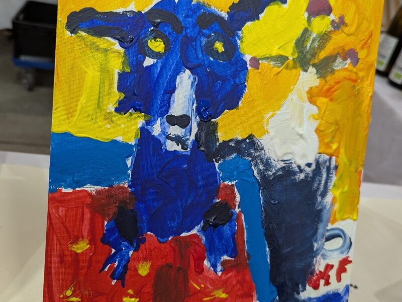 Ein farbenfrohes, abstraktes Gemälde eines blauen Hundes mit gelben Augen auf rotem und blauem Hintergrund, ausgestellt auf einer kleinen Staffelei.