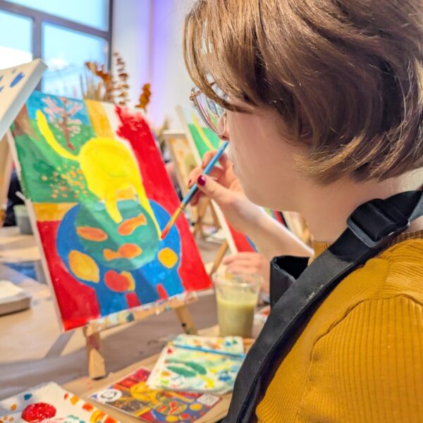 Eine Person mit kurzen Haaren, einer Brille und einem senffarbenen Hemd malt in einem Kunststudio ein buntes abstraktes Bild auf eine Leinwand. Malutensilien liegen auf dem Tisch.
