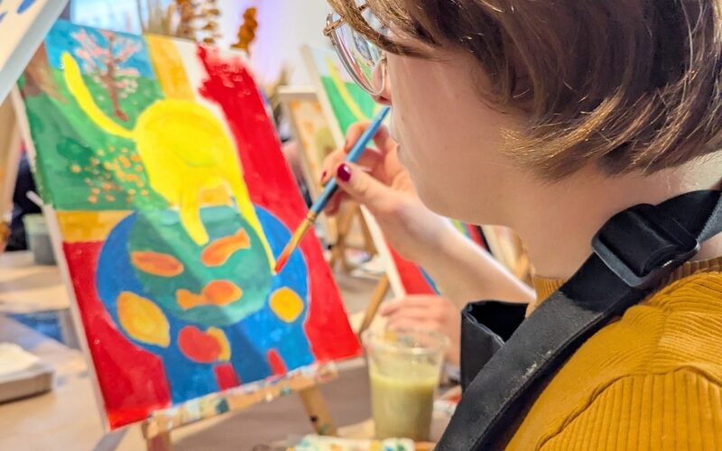 Eine Person mit kurzen Haaren, einer Brille und einem senffarbenen Hemd malt in einem Kunststudio ein buntes abstraktes Bild auf eine Leinwand. Malutensilien liegen auf dem Tisch.