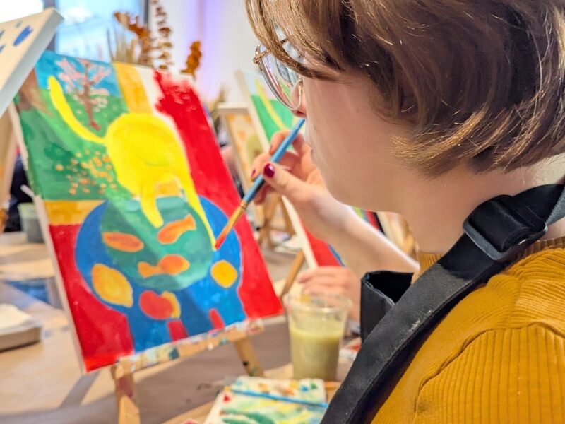 Eine Person mit kurzen Haaren, einer Brille und einem senffarbenen Hemd malt in einem Kunststudio ein buntes abstraktes Bild auf eine Leinwand. Malutensilien liegen auf dem Tisch.