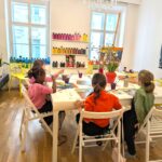 Vier Kinder sitzen um einen Tisch in einem Kunstunterrichtsraum und malen mit Pinseln. Im Hintergrund sind Regale mit bunten Farbflaschen und Kunstzubehör zu sehen.