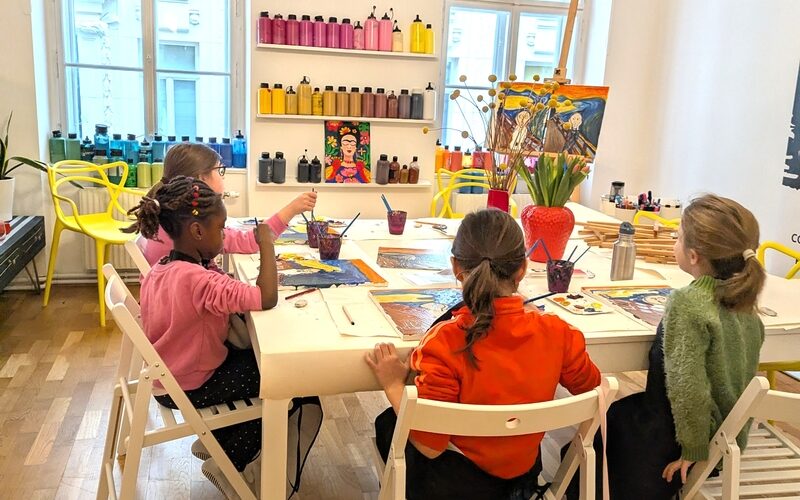 Vier Kinder sitzen um einen Tisch in einem Kunstunterrichtsraum und malen mit Pinseln. Im Hintergrund sind Regale mit bunten Farbflaschen und Kunstzubehör zu sehen.