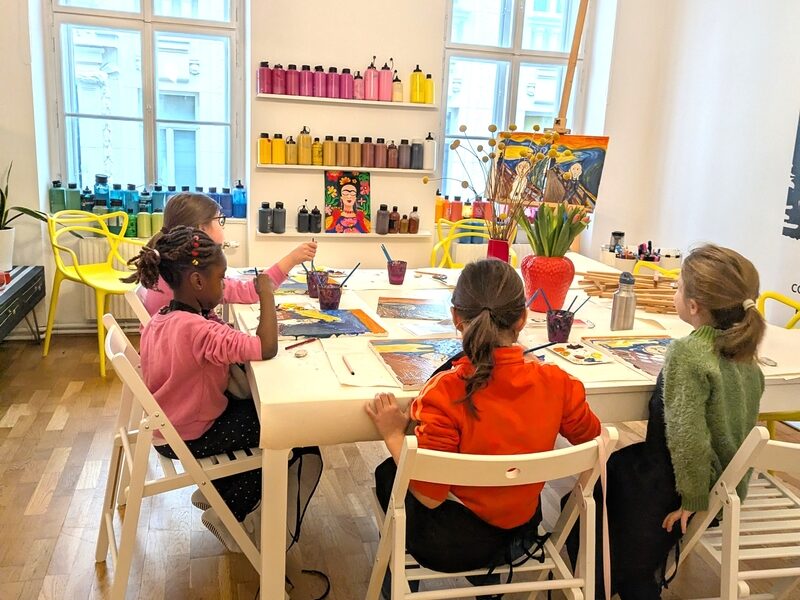 Vier Kinder sitzen um einen Tisch in einem Kunstunterrichtsraum und malen mit Pinseln. Im Hintergrund sind Regale mit bunten Farbflaschen und Kunstzubehör zu sehen.
