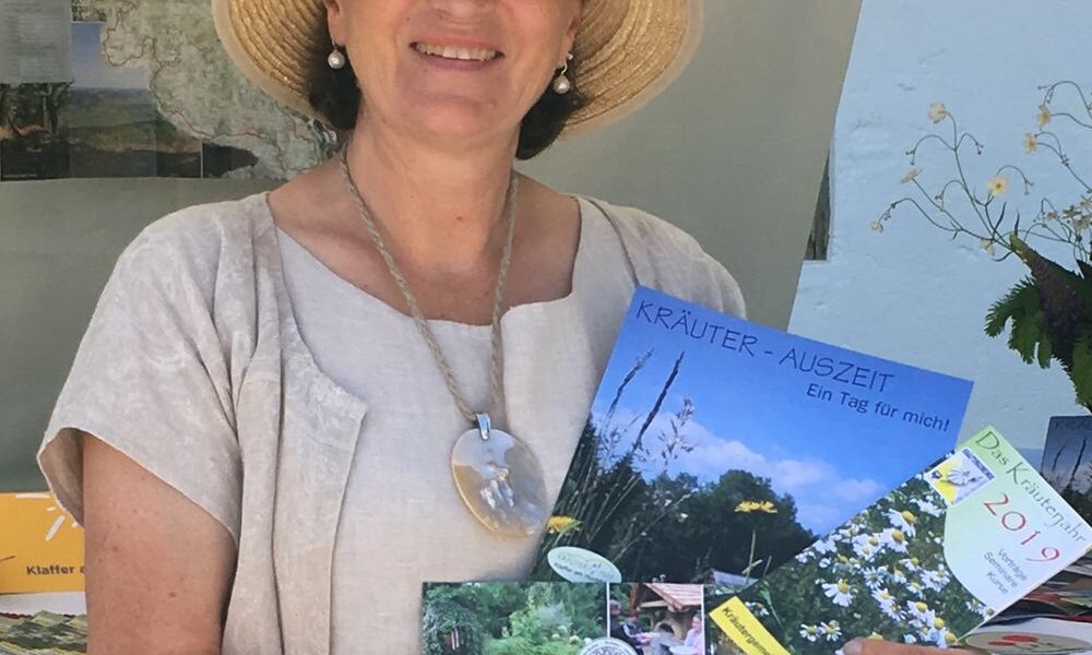 Eine Frau mit Hut und beiger Kleidung hält an einem Informationsstand im Freien Broschüren über Kräuter und Gärten in der Hand.
