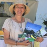 Eine Frau mit Hut und beiger Kleidung hält an einem Informationsstand im Freien Broschüren über Kräuter und Gärten in der Hand.