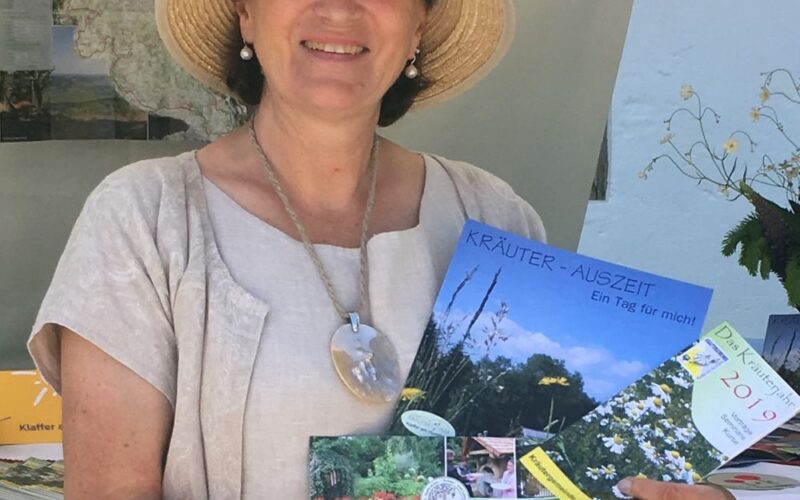 Eine Frau mit Hut und beiger Kleidung hält an einem Informationsstand im Freien Broschüren über Kräuter und Gärten in der Hand.