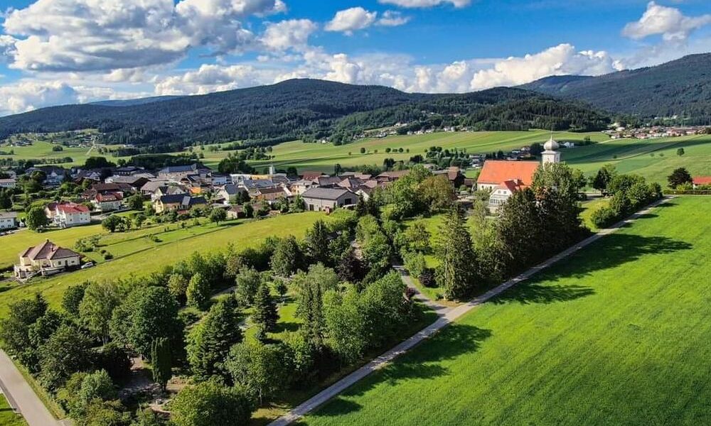 Luftaufnahme eines kleinen Dorfes, umgeben von grünen Feldern, Bäumen und fernen Hügeln unter einem blauen Himmel mit vereinzelten Wolken.