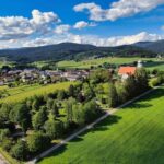 Luftaufnahme eines kleinen Dorfes, umgeben von grünen Feldern, Bäumen und fernen Hügeln unter einem blauen Himmel mit vereinzelten Wolken.