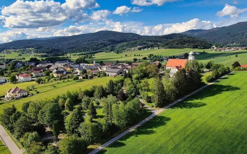 Luftaufnahme eines kleinen Dorfes, umgeben von grünen Feldern, Bäumen und fernen Hügeln unter einem blauen Himmel mit vereinzelten Wolken.