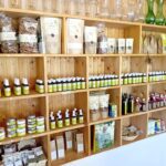 In Holzregalen werden verschiedene Naturprodukte wie Gläser, Flaschen, Päckchen und getrocknete Kräuter in einem Laden ausgestellt.