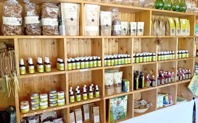 In Holzregalen werden verschiedene Naturprodukte wie Gläser, Flaschen, Päckchen und getrocknete Kräuter in einem Laden ausgestellt.