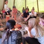 Eine Gruppe von Menschen, meist Kinder, sitzt und steht um ein Lagerfeuer in einem großen Zelt und röstet Essen auf Stöcken über den Flammen.