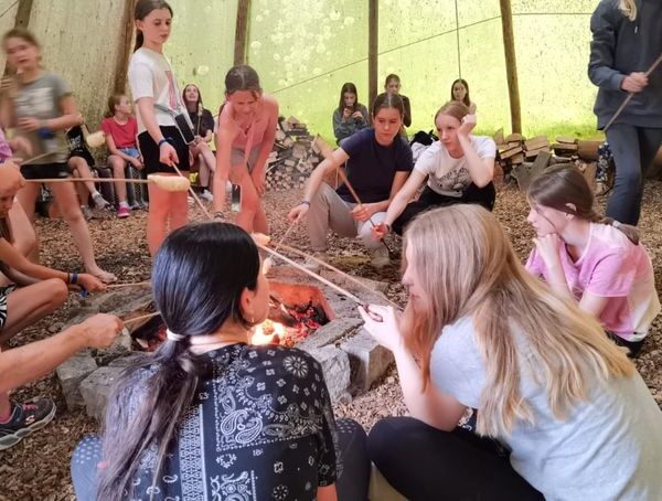 Eine Gruppe von Menschen, meist Kinder, sitzt und steht um ein Lagerfeuer in einem großen Zelt und röstet Essen auf Stöcken über den Flammen.