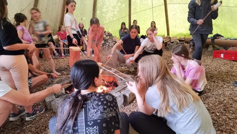 Eine Gruppe von Menschen, meist Kinder, sitzt und steht um ein Lagerfeuer in einem großen Zelt und röstet Essen auf Stöcken über den Flammen.