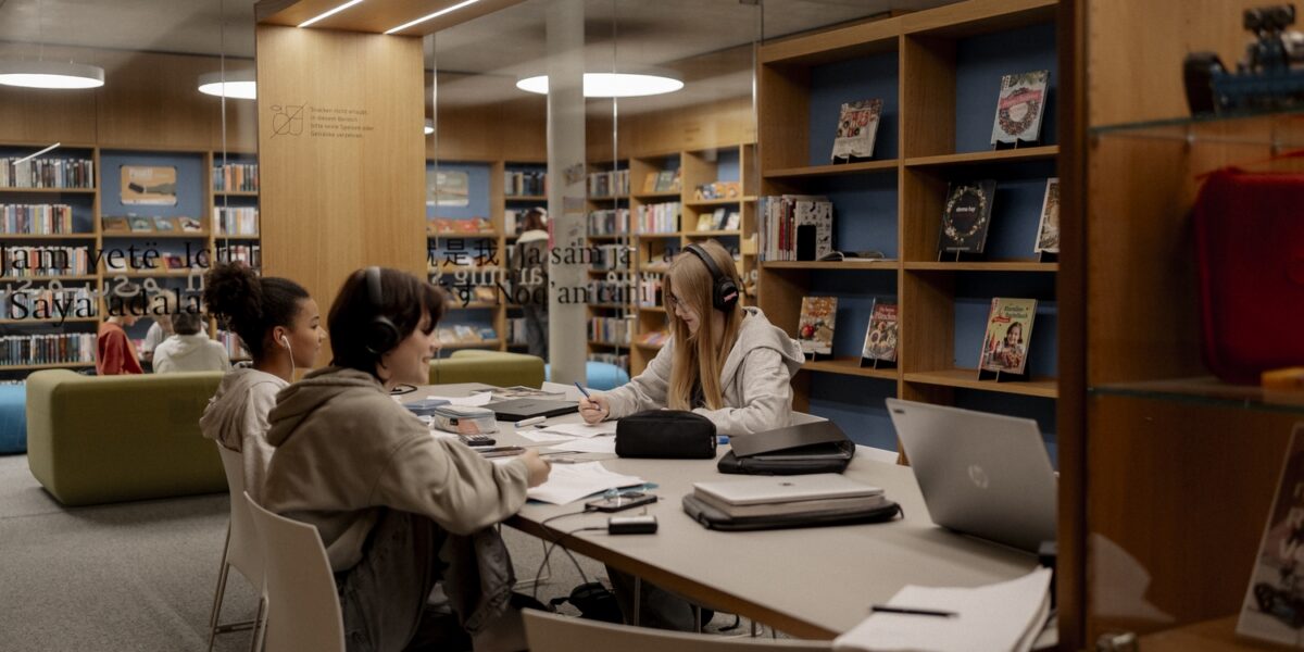 Drei Personen sitzen an einem Tisch in einer modernen Bibliothek, studieren und arbeiten an Laptops und Papieren, umgeben von Bücherregalen und Lehrmaterial.