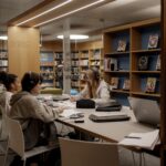 Drei Personen sitzen an einem Tisch in einer modernen Bibliothek, studieren und arbeiten an Laptops und Papieren, umgeben von Bücherregalen und Lehrmaterial.