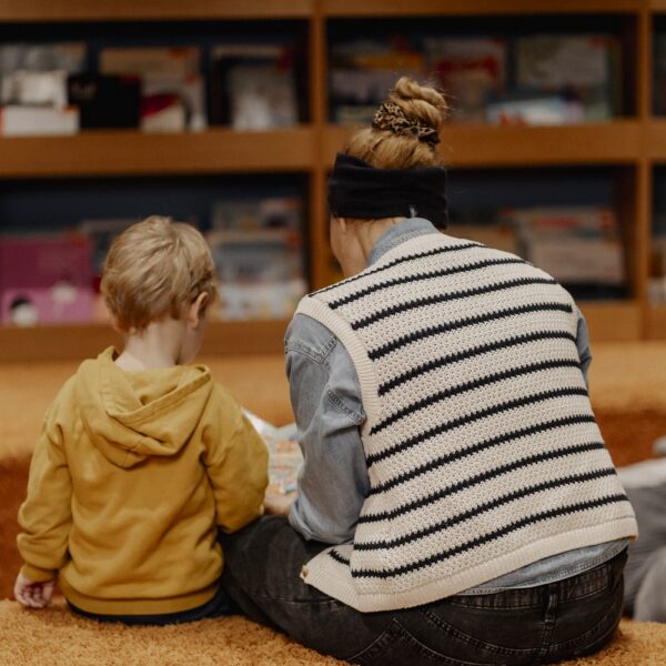 Ein Erwachsener und ein Kind sitzen auf einem Teppichboden, mit dem Gesicht zur Seite, und schauen sich gemeinsam ein Buch vor einem Bücherregal an.