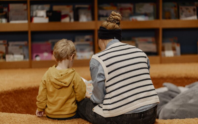Ein Erwachsener und ein Kind sitzen auf einem Teppichboden, mit dem Gesicht zur Seite, und schauen sich gemeinsam ein Buch vor einem Bücherregal an.