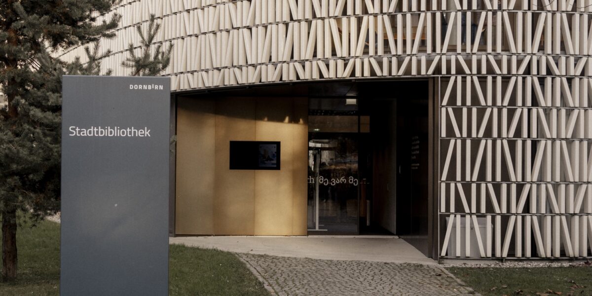 Ein modernes rundes Gebäude mit einer gemusterten Fassade mit der Aufschrift "Stadtbibliothek"; ein Schild und ein Kopfsteinpflasterweg führen zum Eingang.