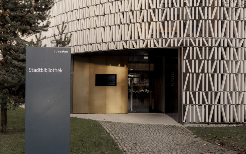 Ein modernes rundes Gebäude mit einer gemusterten Fassade mit der Aufschrift "Stadtbibliothek"; ein Schild und ein Kopfsteinpflasterweg führen zum Eingang.