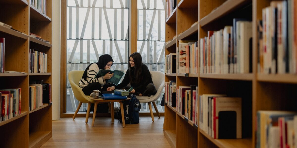 Zwei Personen sitzen auf Stühlen zwischen Bücherregalen in einer Bibliothek, lesen und unterhalten sich, mit großen Fenstern im Hintergrund.