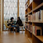 Zwei Personen sitzen auf Stühlen zwischen Bücherregalen in einer Bibliothek, lesen und unterhalten sich, mit großen Fenstern im Hintergrund.