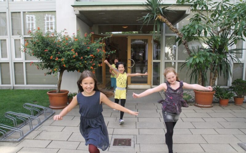 Drei junge Mädchen rennen und lächeln vor einem Gebäude mit großen Fenstern, umgeben von Topfpflanzen und Grünpflanzen auf einem gepflasterten Gehweg.