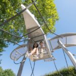 Zwei Kinder sitzen in einem an Seilen aufgehängten Spielplatz in Form eines Metallflugzeugs, mit Bäumen und einem klaren blauen Himmel im Hintergrund.